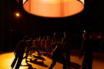 Cloaked in darkness, standing against a dimmed light, dancers are lined up as some move in and out of the diagonal they are standing in.