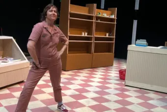 Photo of Lisa Damour standing in a pale red boiler suit on a checkered linoleum floor part of the grocery store set of her play frozen section