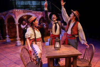 Three undergraduate actors perform in a play, cheering and smiling while standing around a table