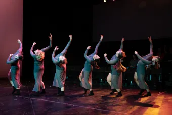 A group of six dancers perform on stage, their arms reaching upward as they lean backward