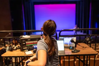 An undergraduate stage manager sits behind a tech table, looking ahead at an empty stage