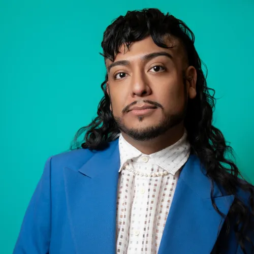 Headshot of a man in a bright blue suit, long curly black hair and a teal backdrop