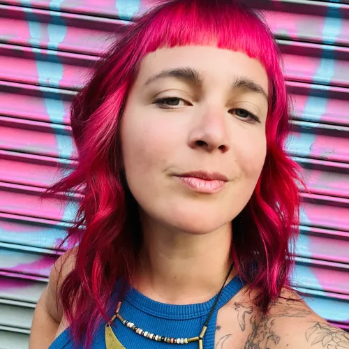 A woman with pink hair and bangs stands in front of a pink metal background
