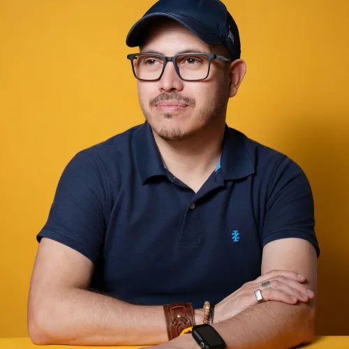 A man in a navy shirt wearing a navy baseball caps sits in front of a yellow background