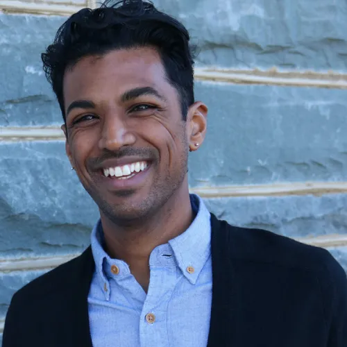 A man in a blue collared shirt with a black sweater smiles