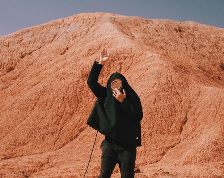 a person in all black stands in front of a mountain of red rock, holding a round piece of wood in front of their face and lifting their right hand