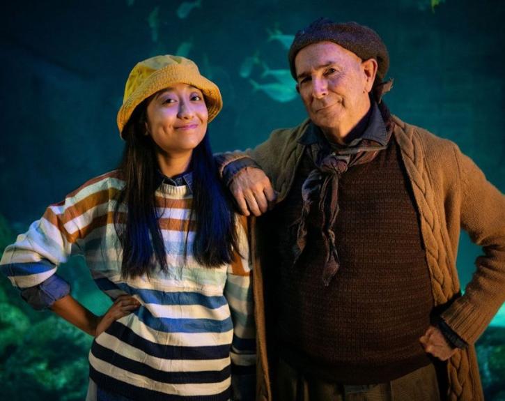 A woman wearing a striped nautical t shirt and a fisherman's cap smiles alongside a gruff but kind mariner