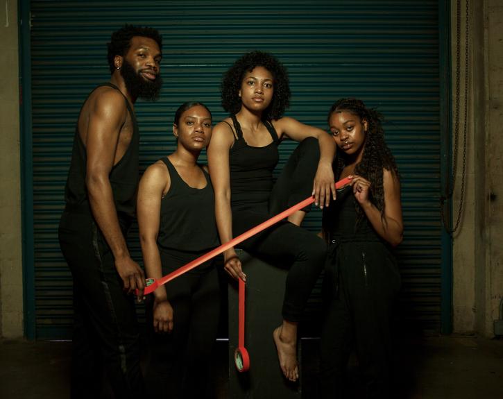 four student artists in black pants and tank tops pose together, holding red tape