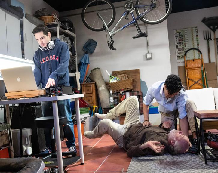 Three actors perform in TOROS, one standing behind a laptop, in what looks like a garage studio space