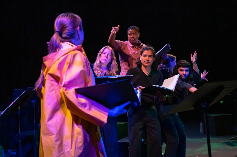 actress holding a black binder looks at a group of actors huddled together and looking ominously back at her