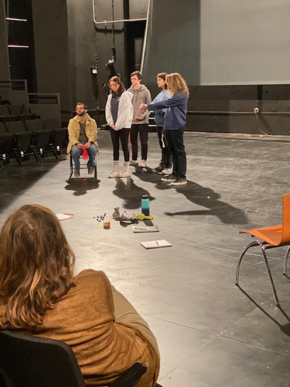 five students perform on stage, one sitting and the others standing, while Rude Mechs members watch and provide guidance