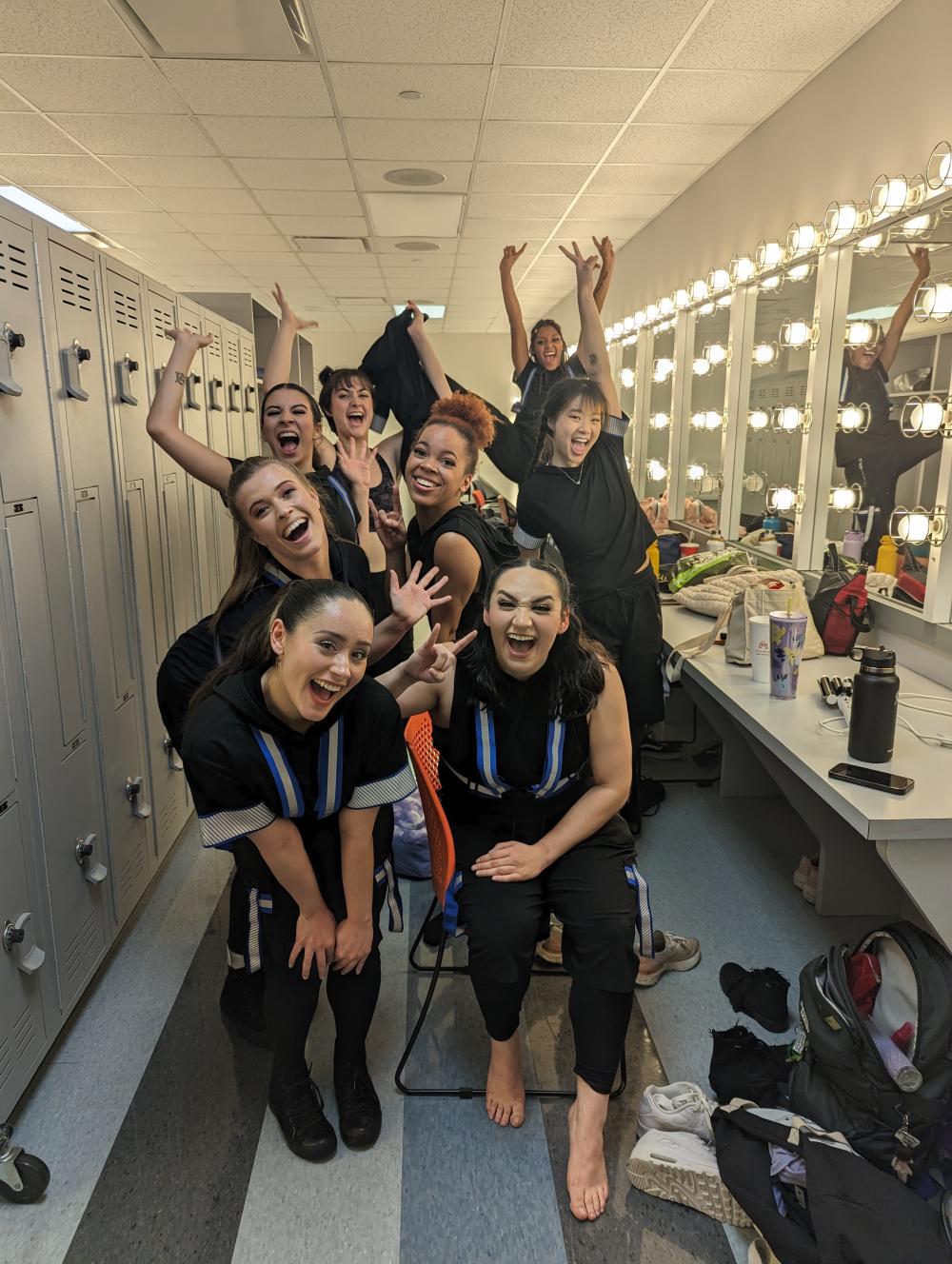 eight dancers pose with their arms raised excitedly in a dressing room with mirrors on one side and lockers on the other