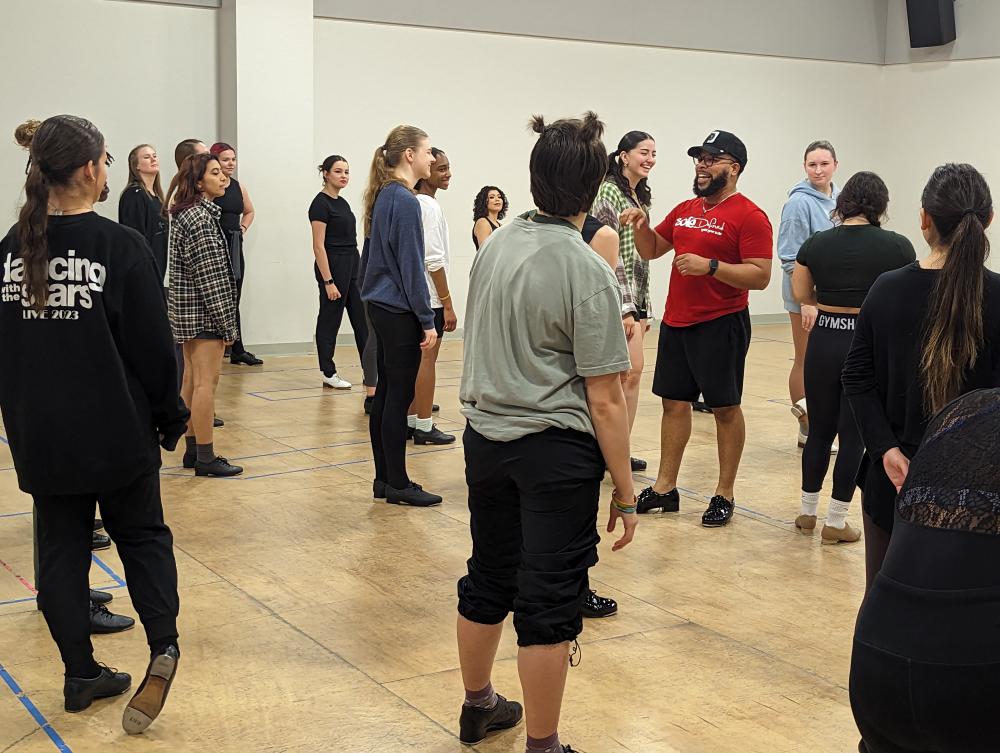 around 17 dancers stand spaced out in a dance studio, watching Ryan K. Johnson as he leads a tap workshop