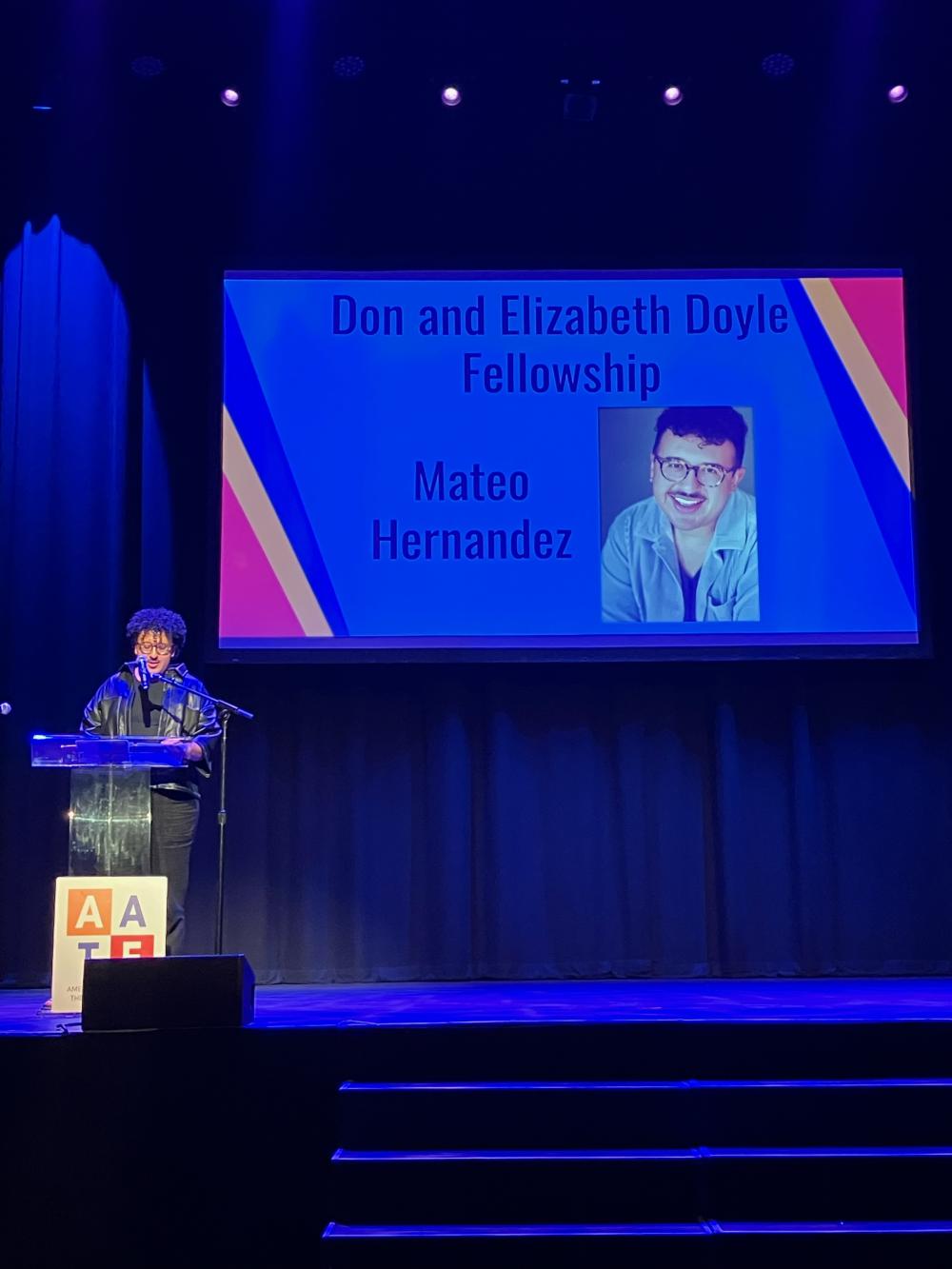 Graduate student Mateo Hernandez standing behind a podium on stage, accepting the Don and Elizabeth Doyle Fellowship