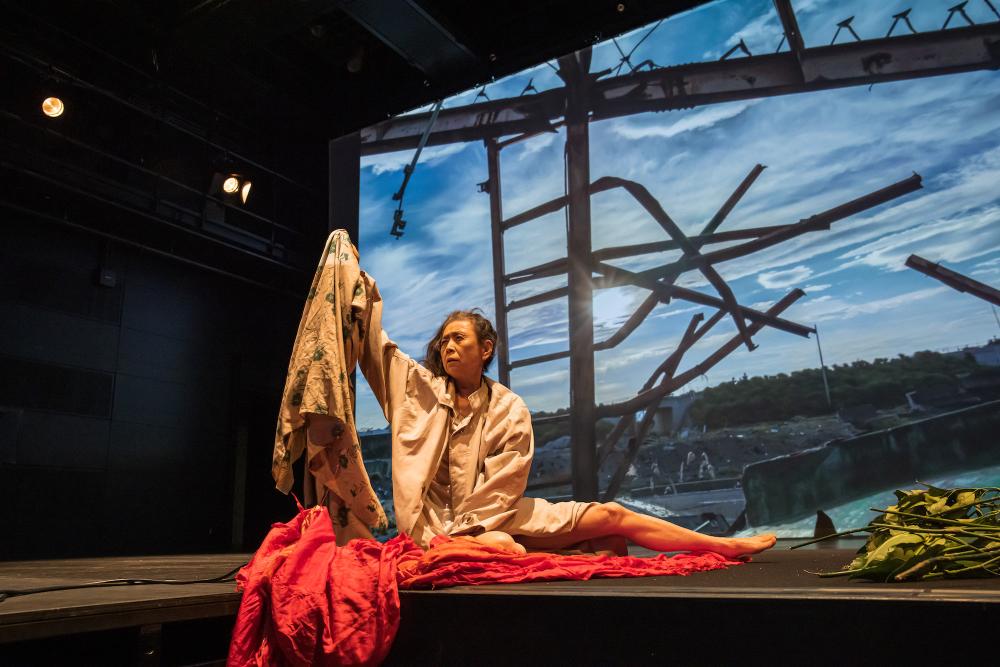 Eiko Otake performing in a movement piece, holding up a patterned fabric, with red fabric in front of her and a projection of the sky and gnarled metal pipes behind her