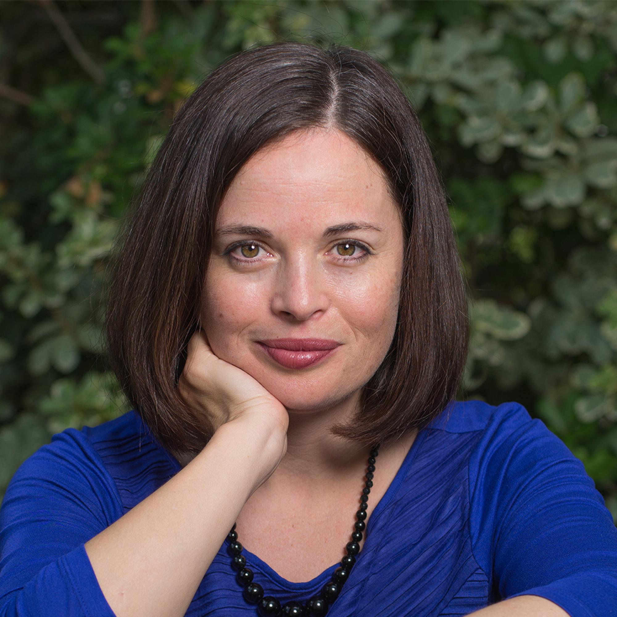 woman with shoulder length brown hair smiles slightly, with her chin propped up on her right hand