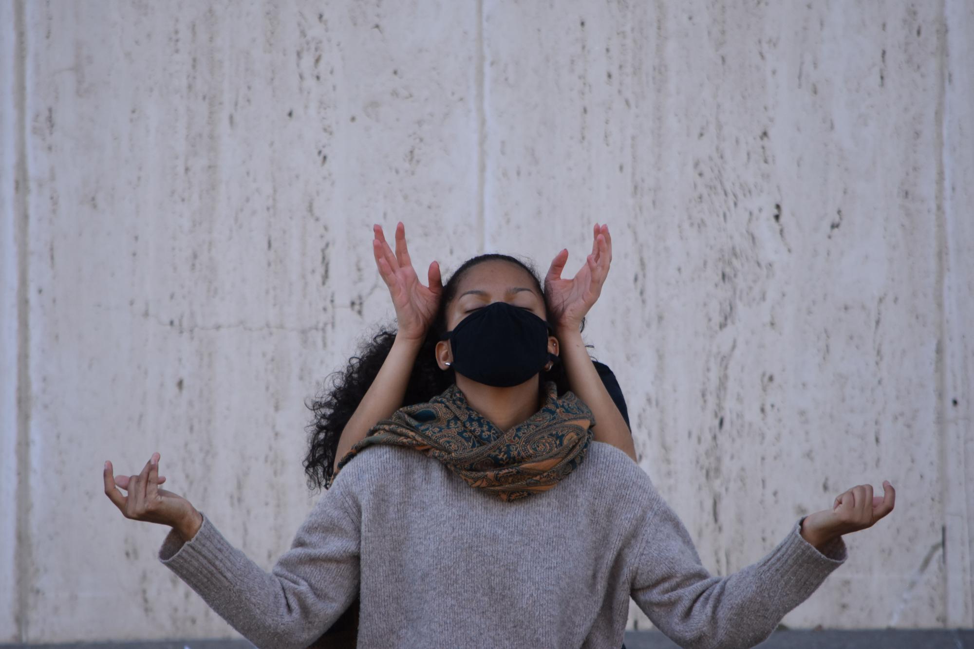 dancer in a drown sweater and black mask holds her arms out and looks up, while another dancer behind her places her hands beside her head