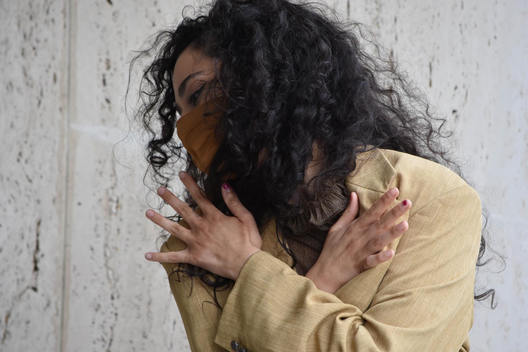 dancer with dark, curly hair, a mask and a tan blazer looks down and crosses her hands over her chest