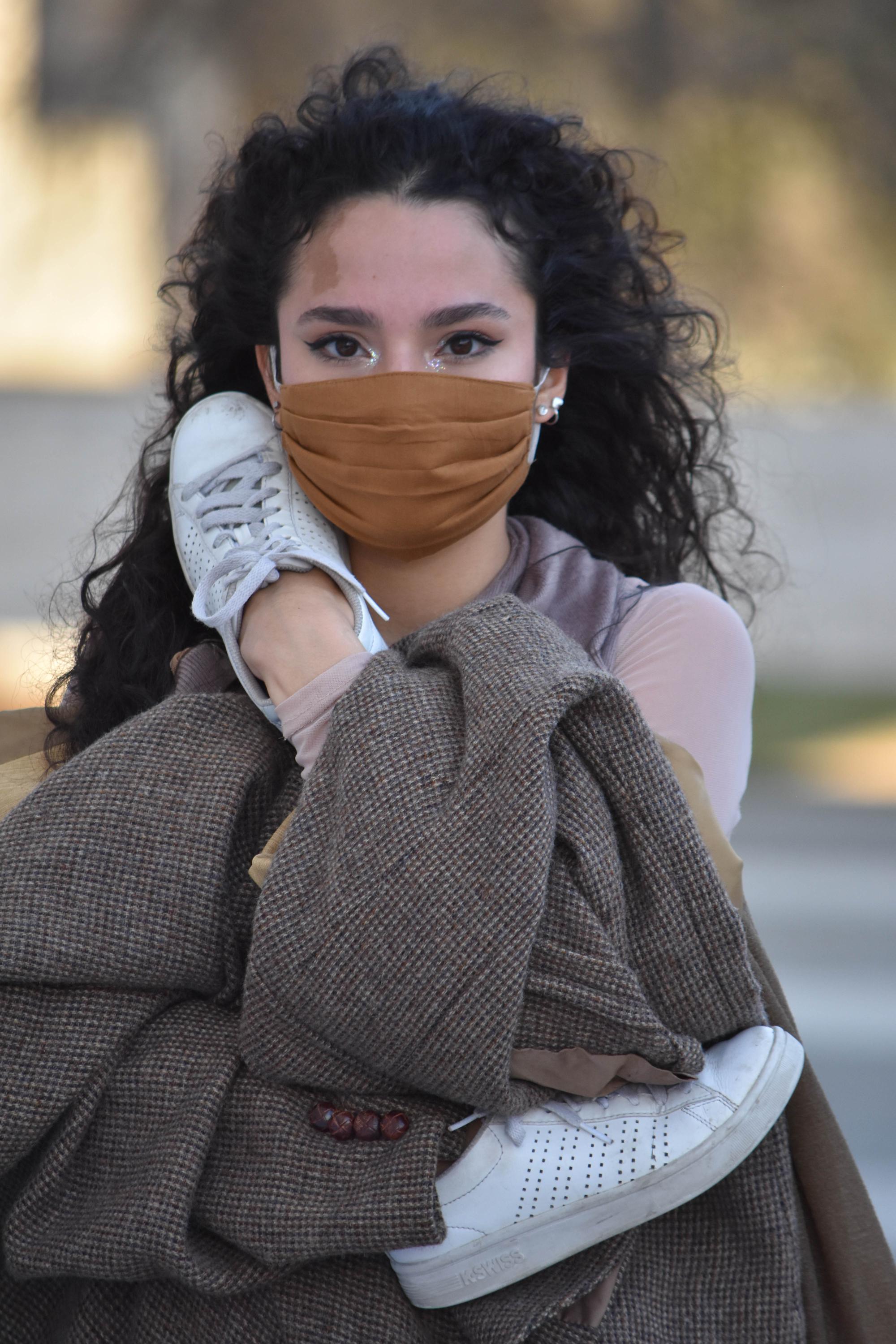 dancer with dark, curly hair and a mask holds a blazer and her tennis shoes across her body