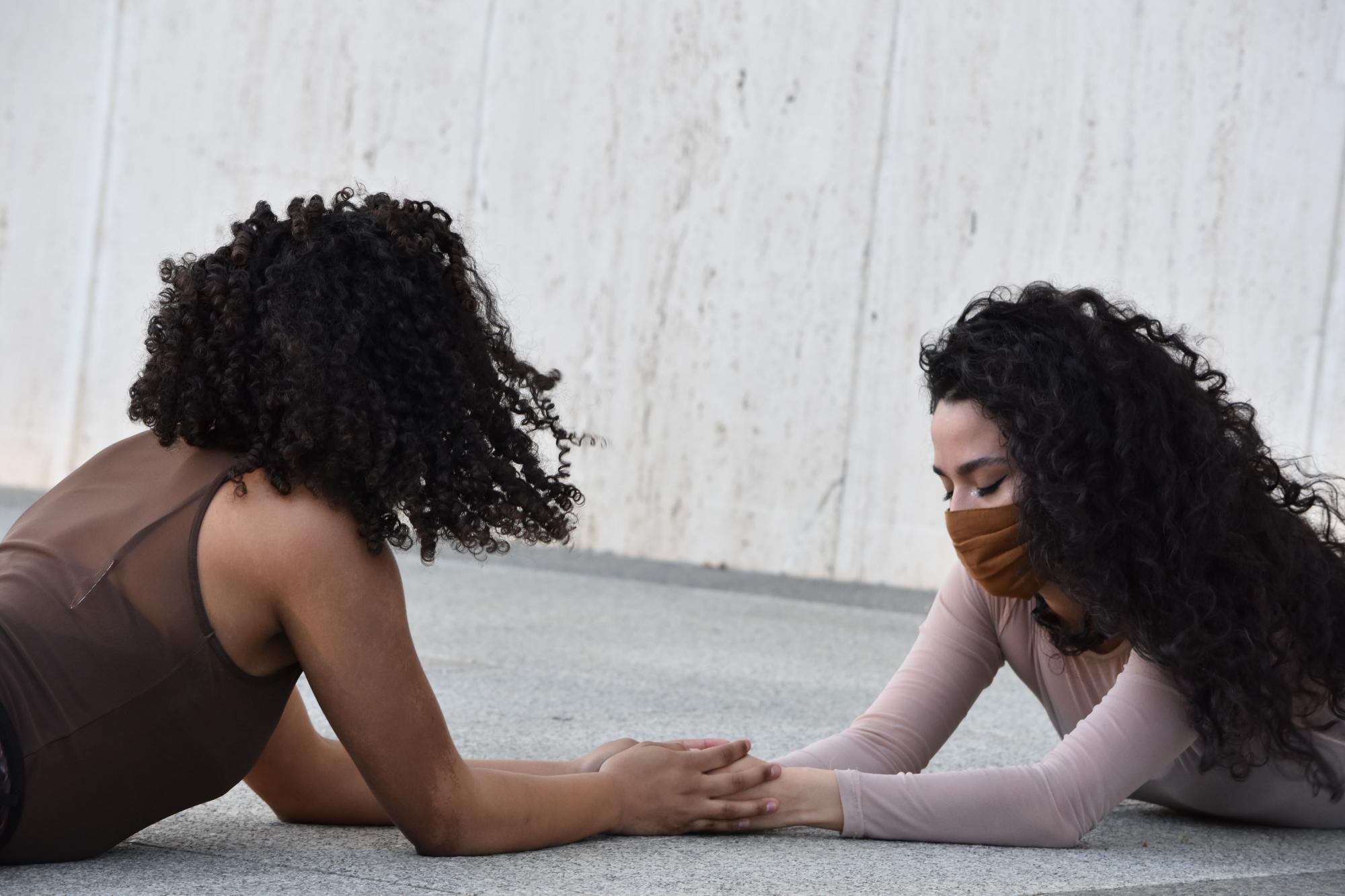 two women with curly black hair lie on their stomaches facing each other and holding hands