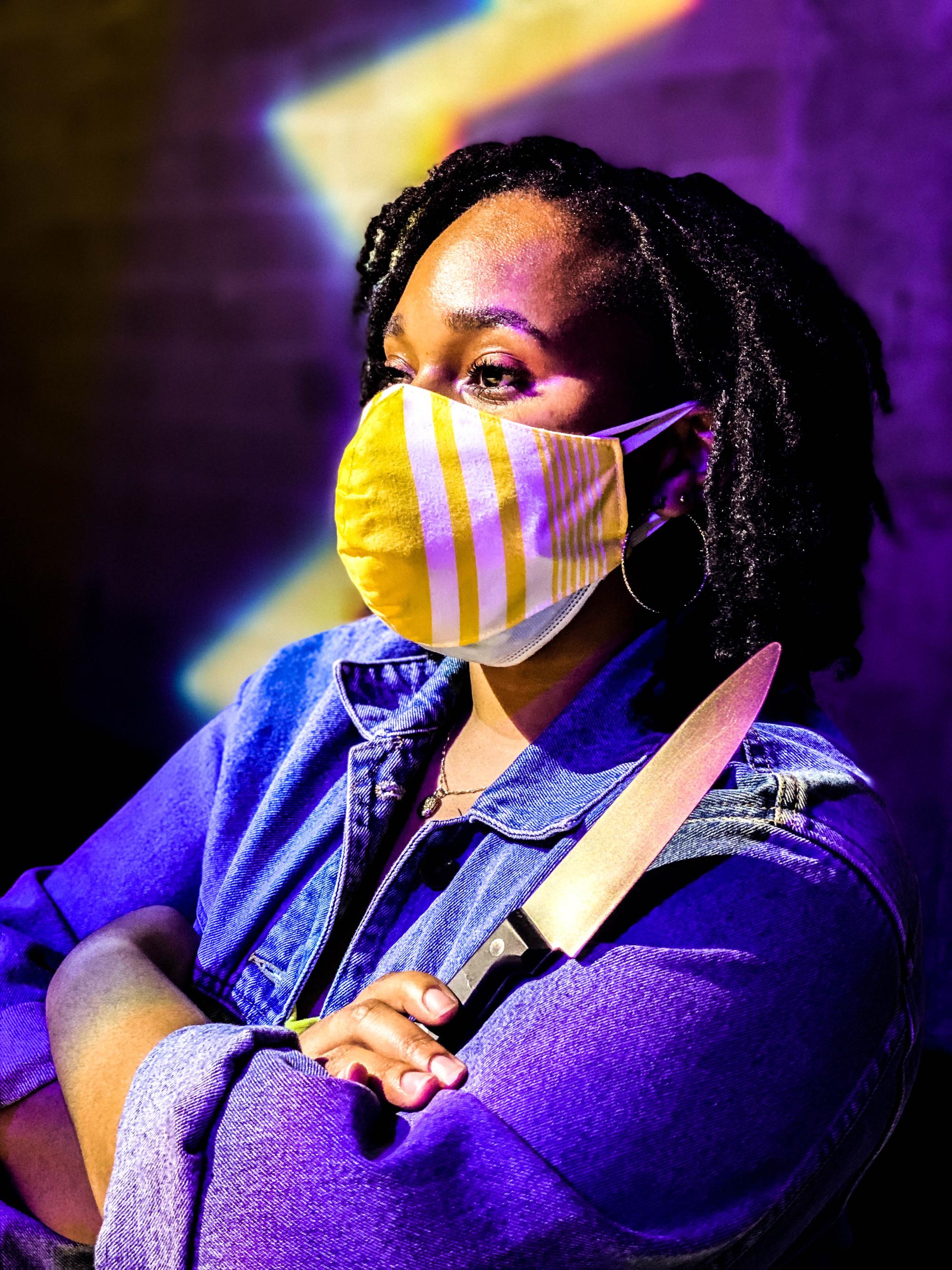 a woman with short dark hair and a yellow mask crosses her arms with a knife in one hand