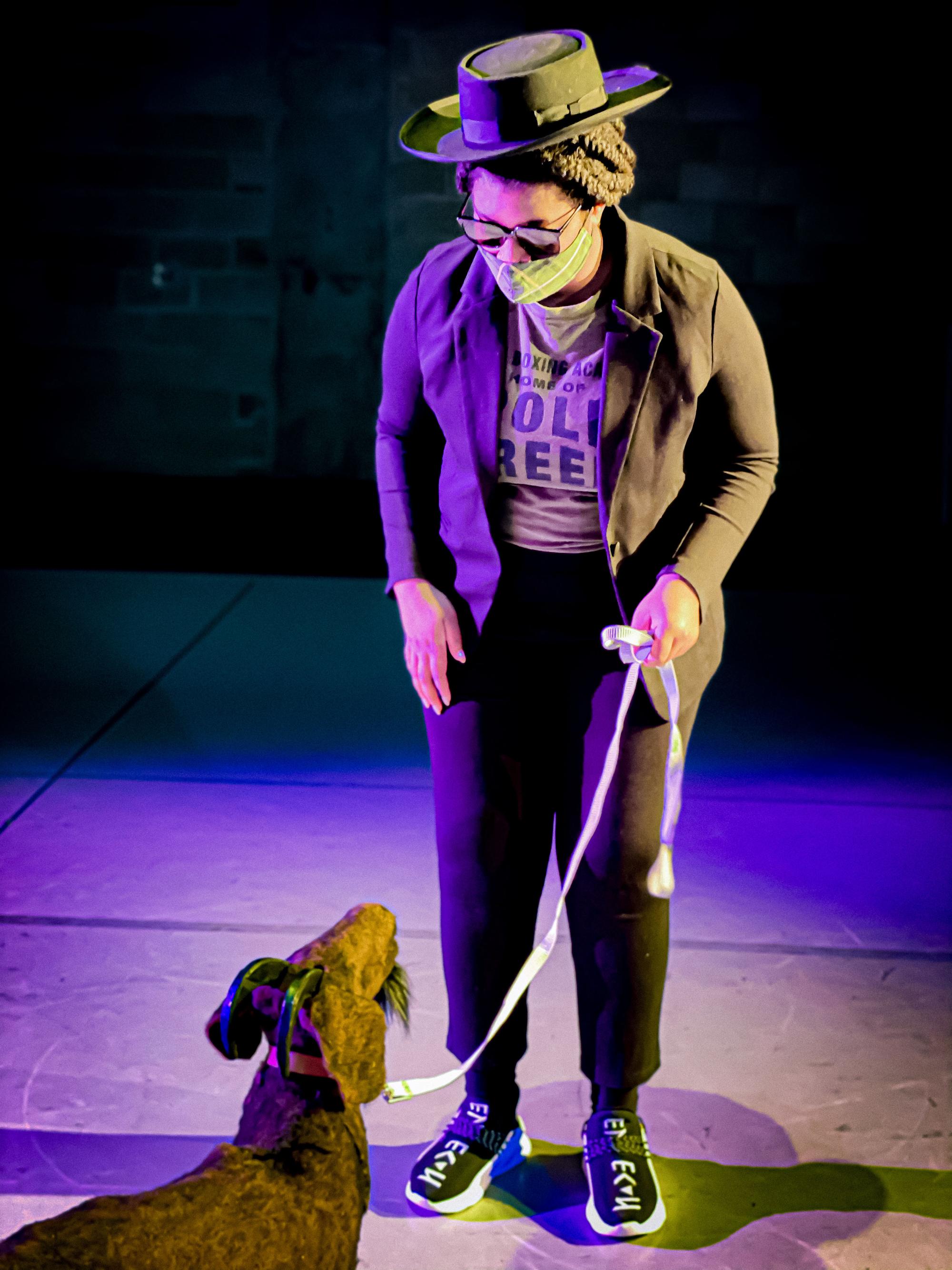 actress in a black hat and sunglasses holds a goat puppet on a leash