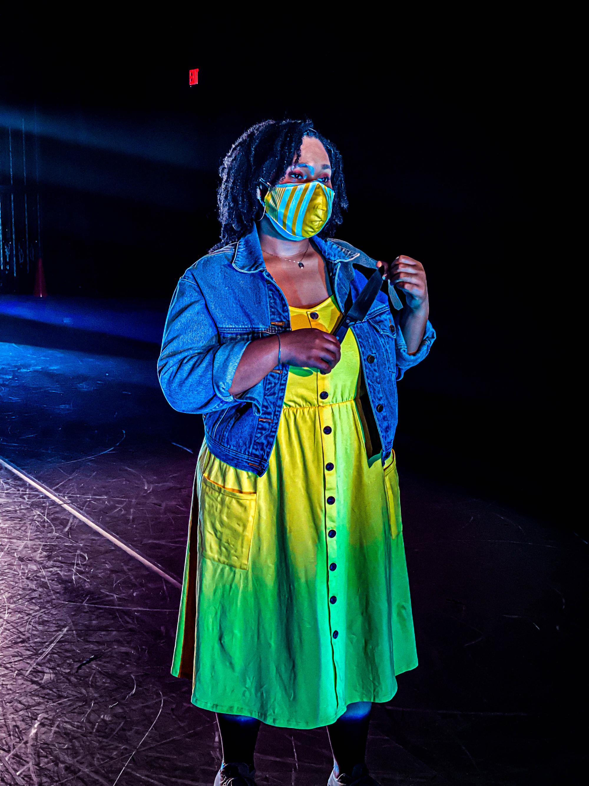 woman in a yellow dress and mask and a blue jean jacket holds a knife