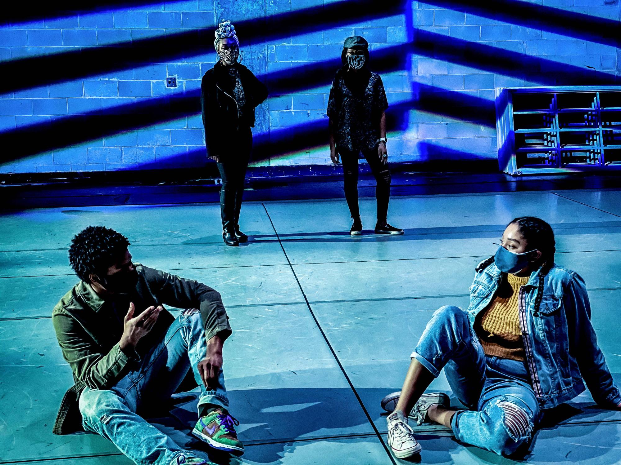 a boy and a girl, both wearing masks, jeans and jackets, sit next to each other and talk, with two women watching them in the background