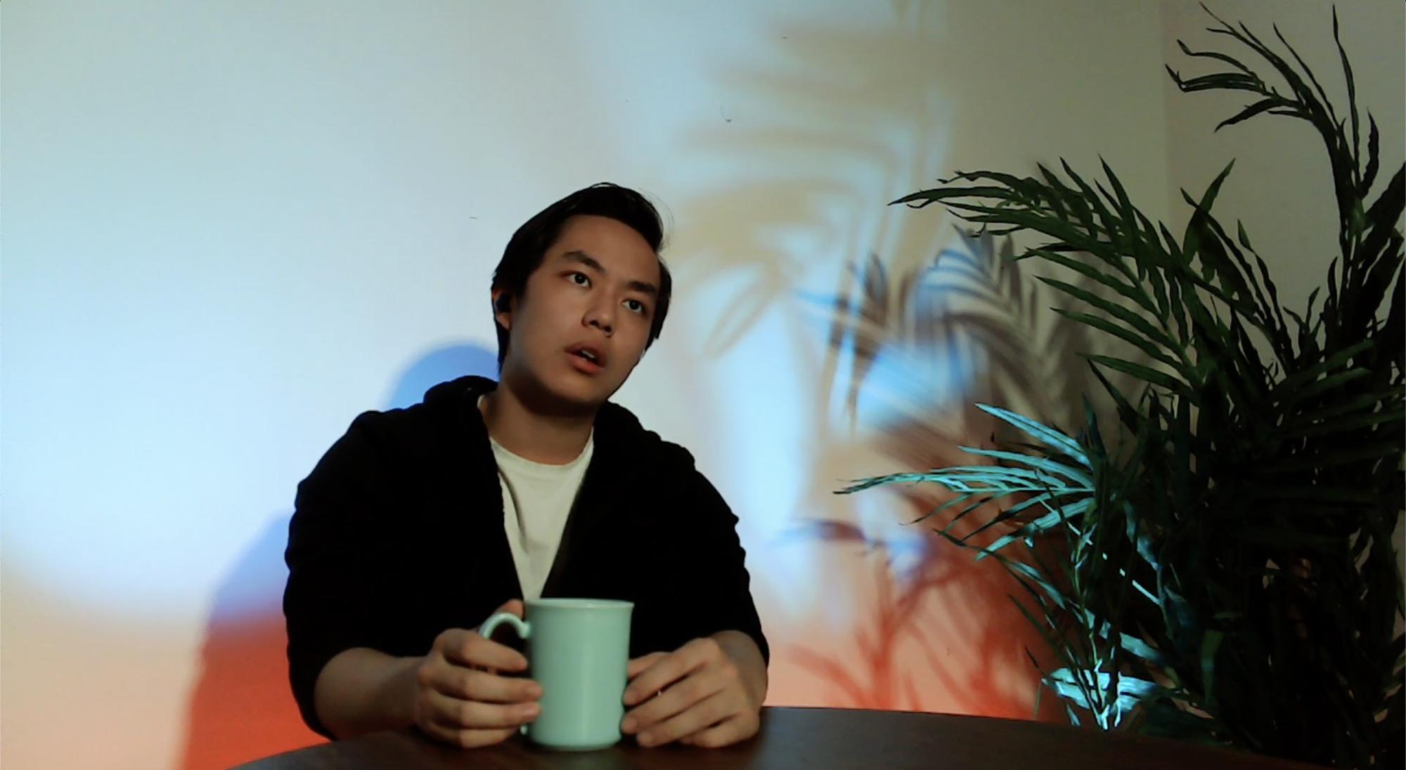 a man with short black hair holding a coffee mug sits at a table, with blue and red light on the wall behind him