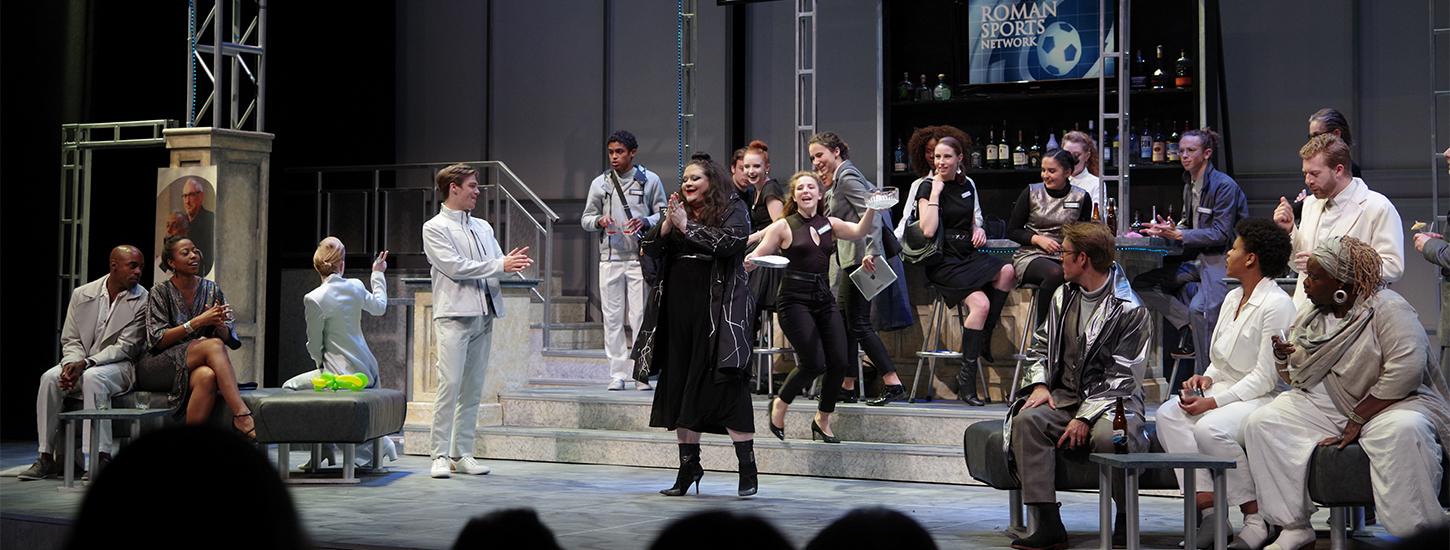 an ensemble of actors on stage, all dressed in shades of white, black or gray with a bar in the background