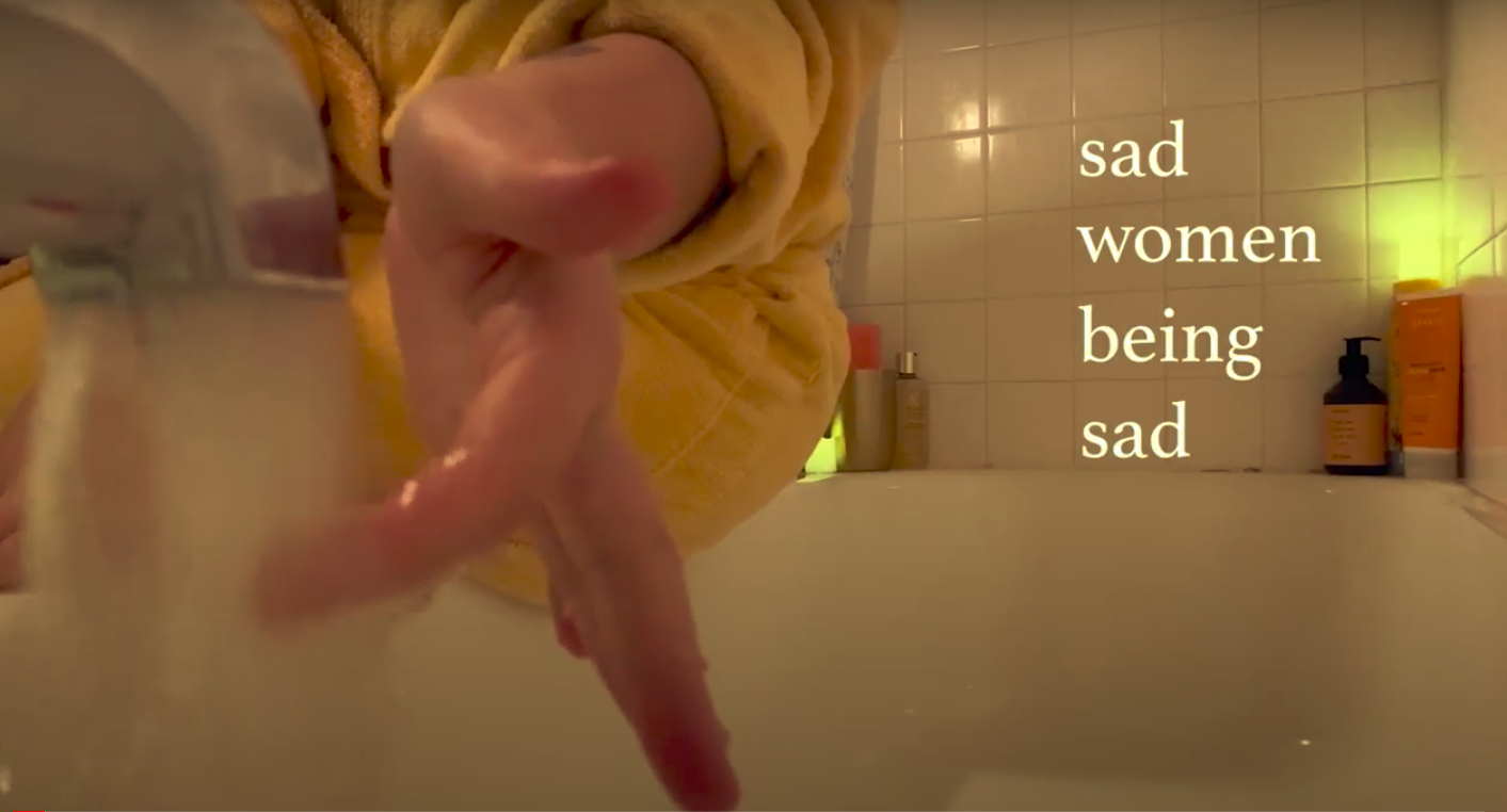 a woman tests the water running from a bathtub faucet with her hand, with the words "sad women being sad" in white on the right of the screen