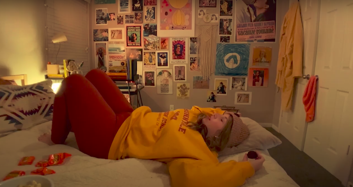 actress in a yellow sweater and beanie lies down on her bed with candy wrappers around her