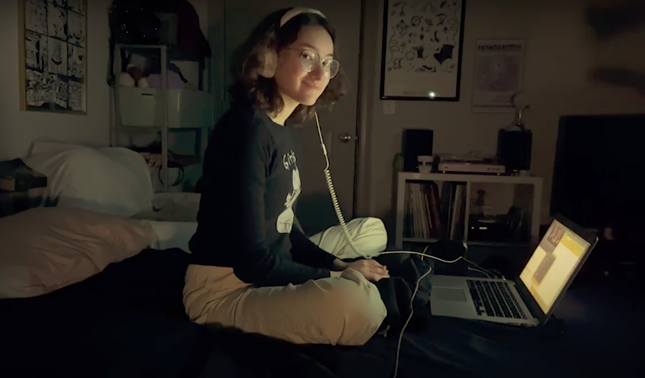 actress wearing headphones sits on her bed in the dark, her face lit up by her laptop screen