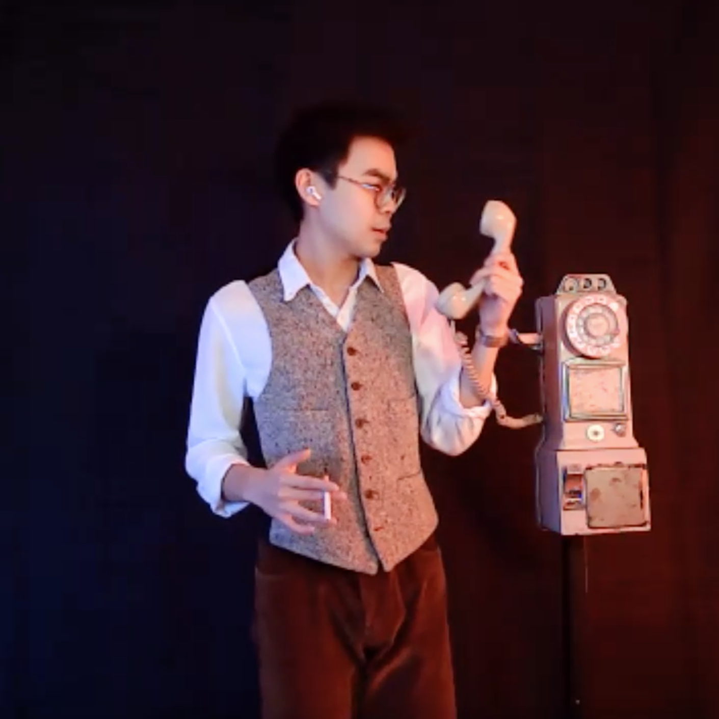 man with glasses and a vest holds a rotary phone in one hand and a cigarette in the other
