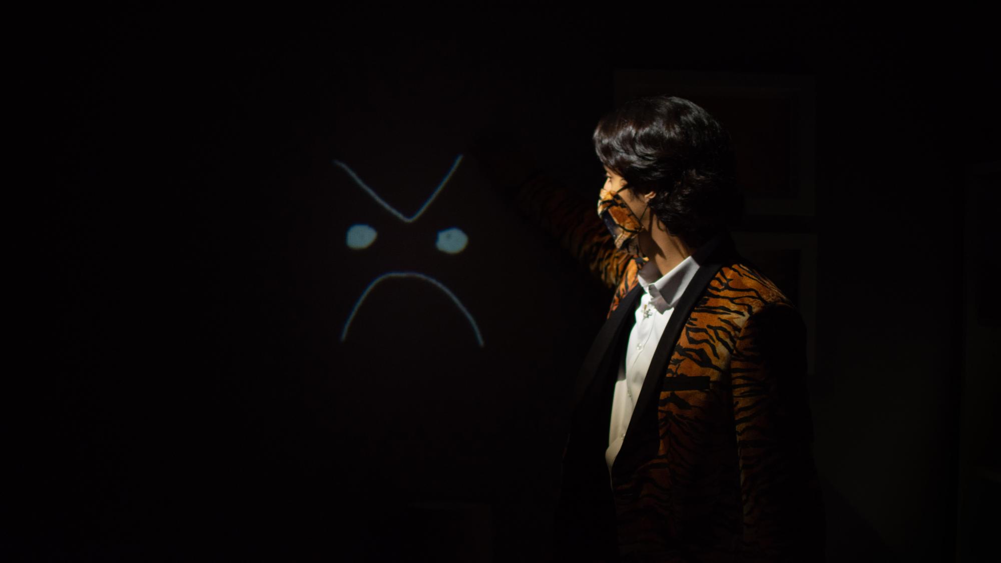 actor in a tiger striped suit coat and mask points at a projection