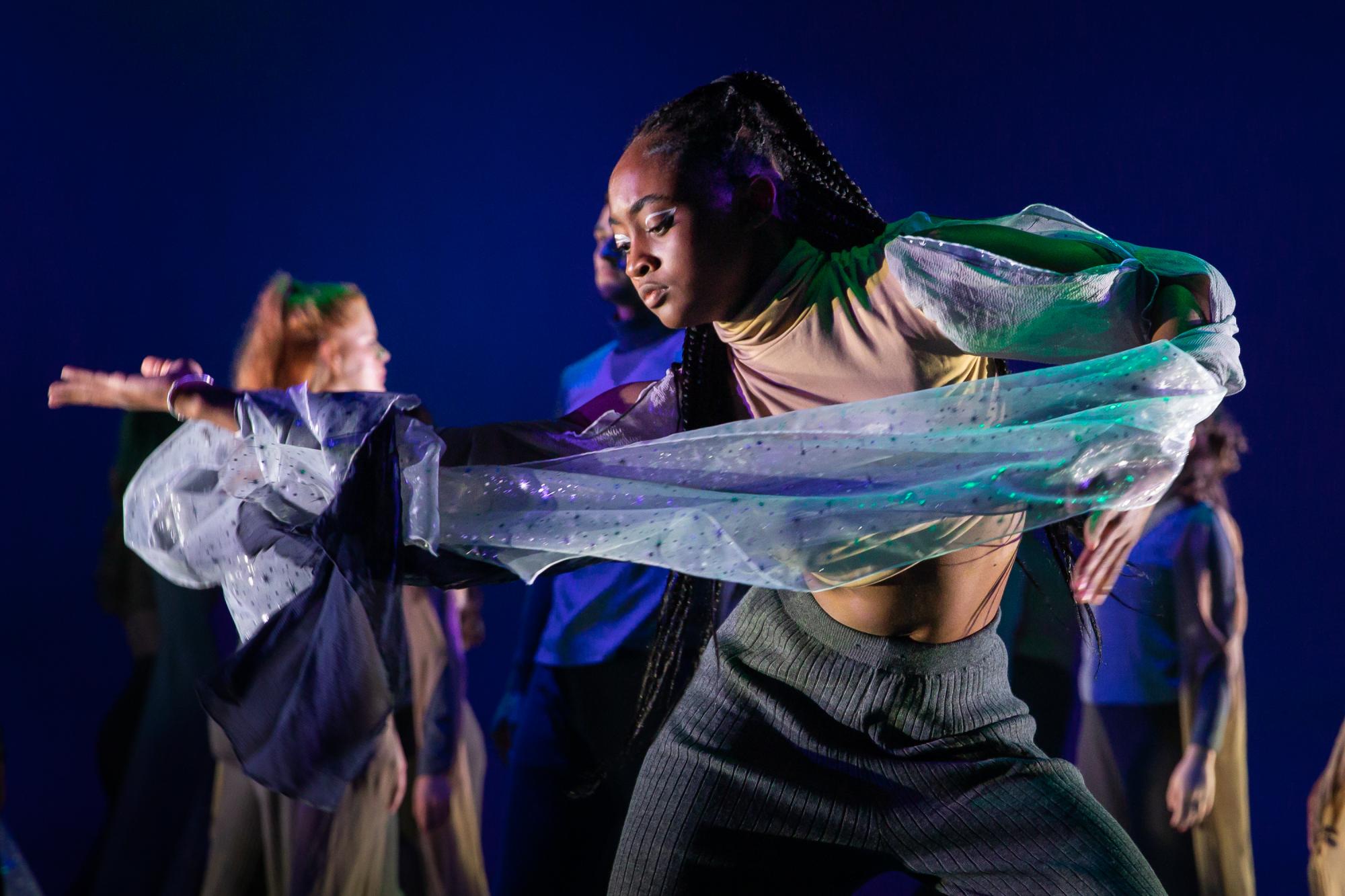 dancer holds her right arm out beside her, the blue fabric of her left sleeve trailing between her two hands