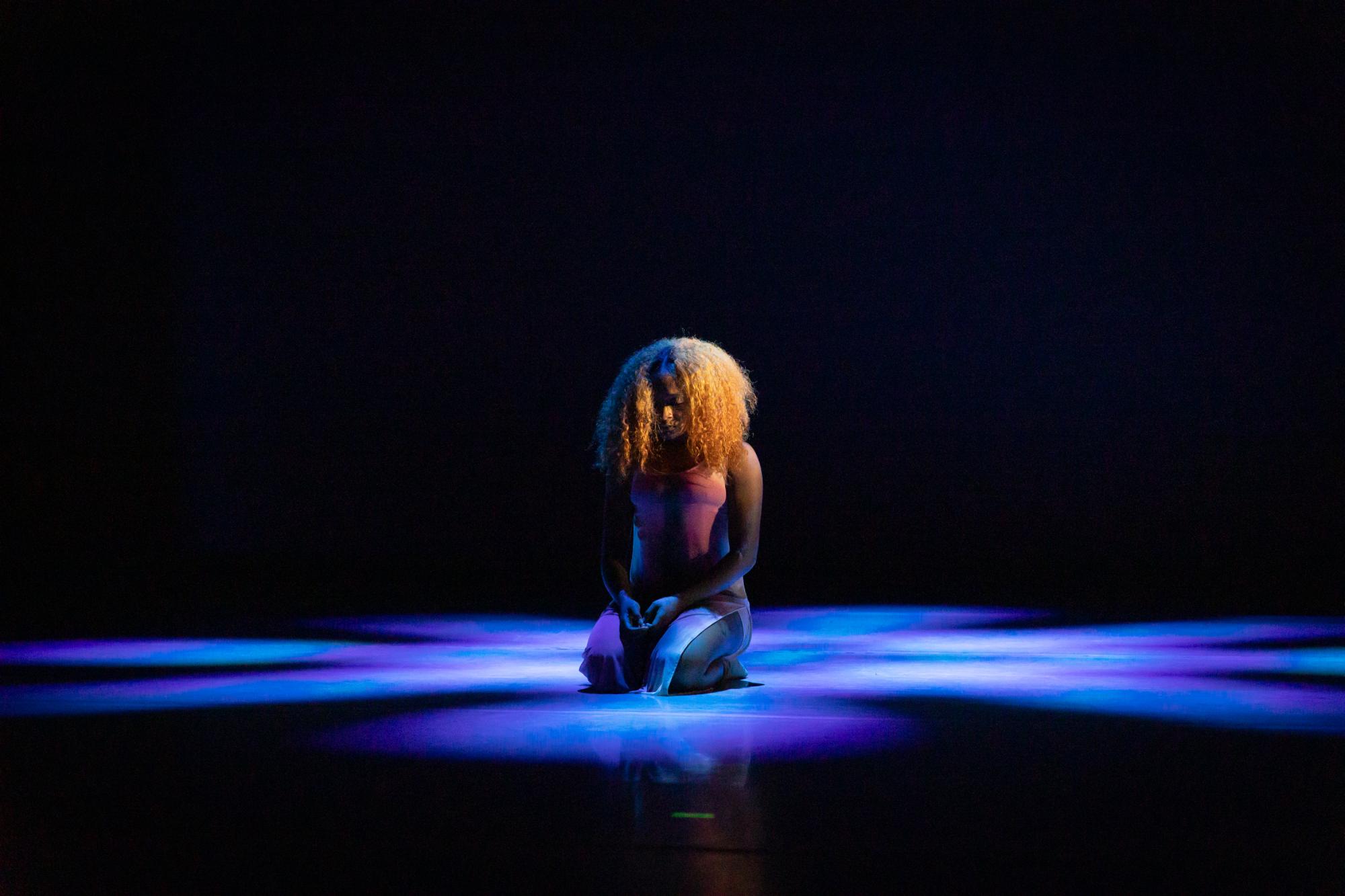 a dancer with curly blonde hair sits on her knees, looking down, with blue light shining on her
