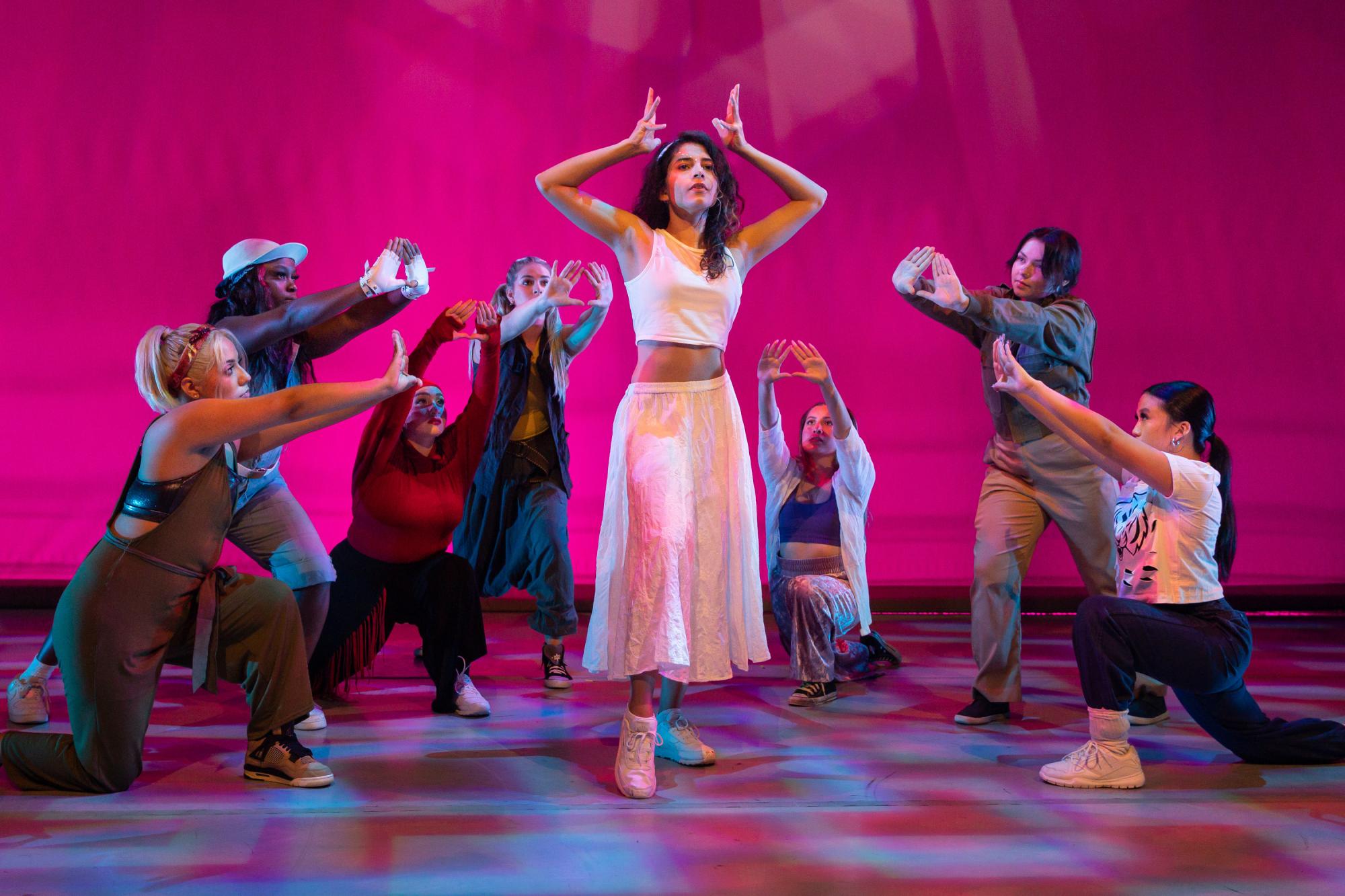 seven dancers form a semi-circle around an eighth dancer, who holds her hands above her head like a crown