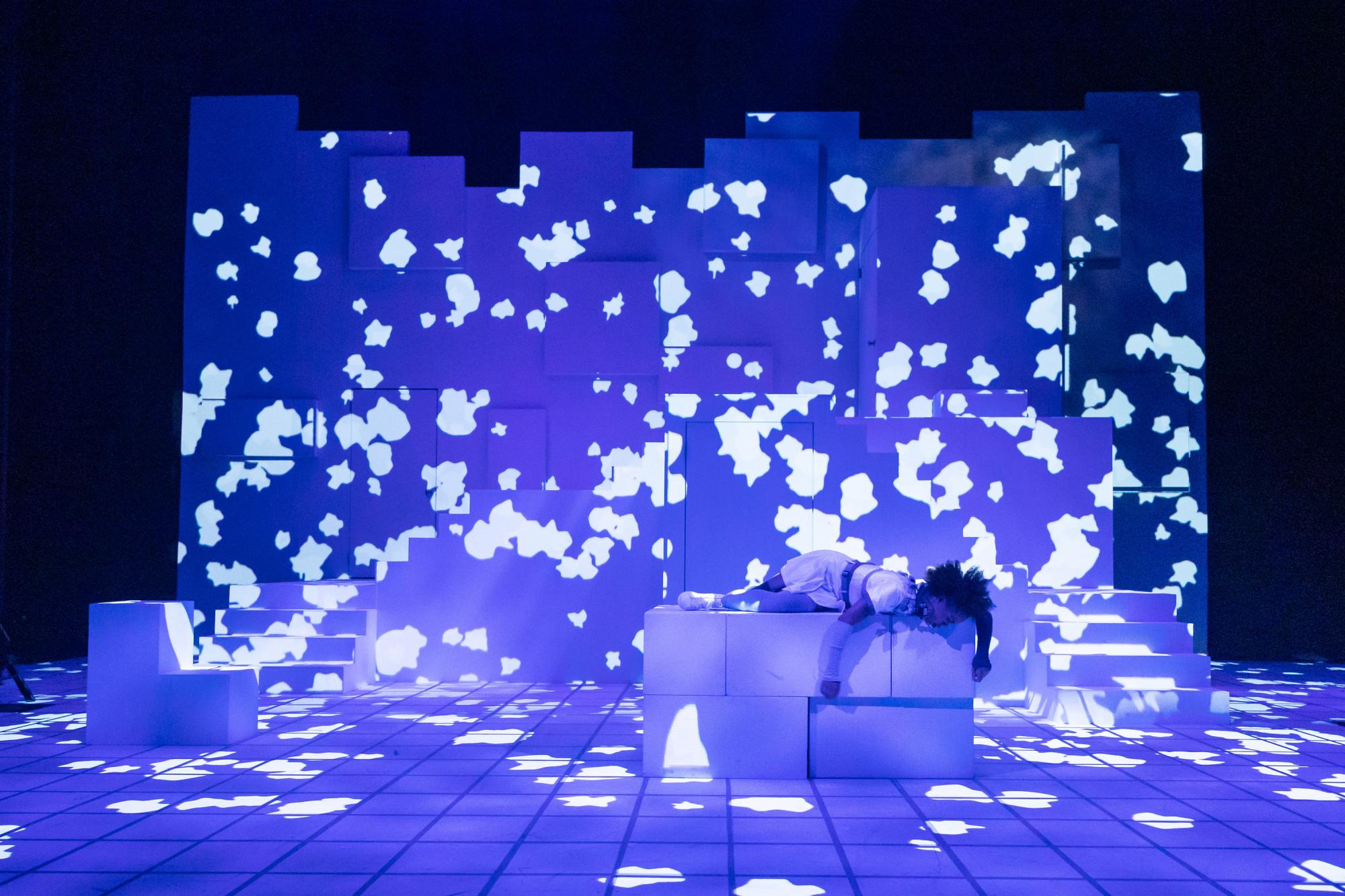 an actress lies on white blocks on stage, with vivid blue and white projections all around her