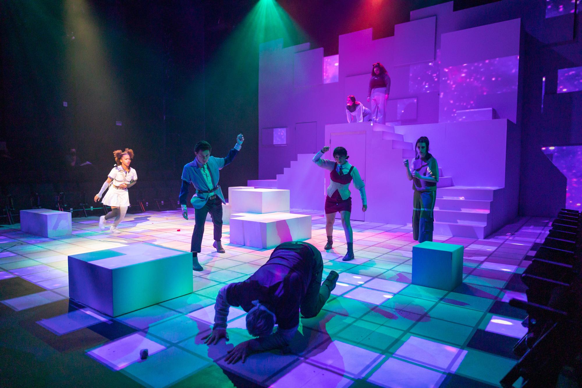 three actors throw things at a fourth actor who's on the ground, with colorful stage lighting around them