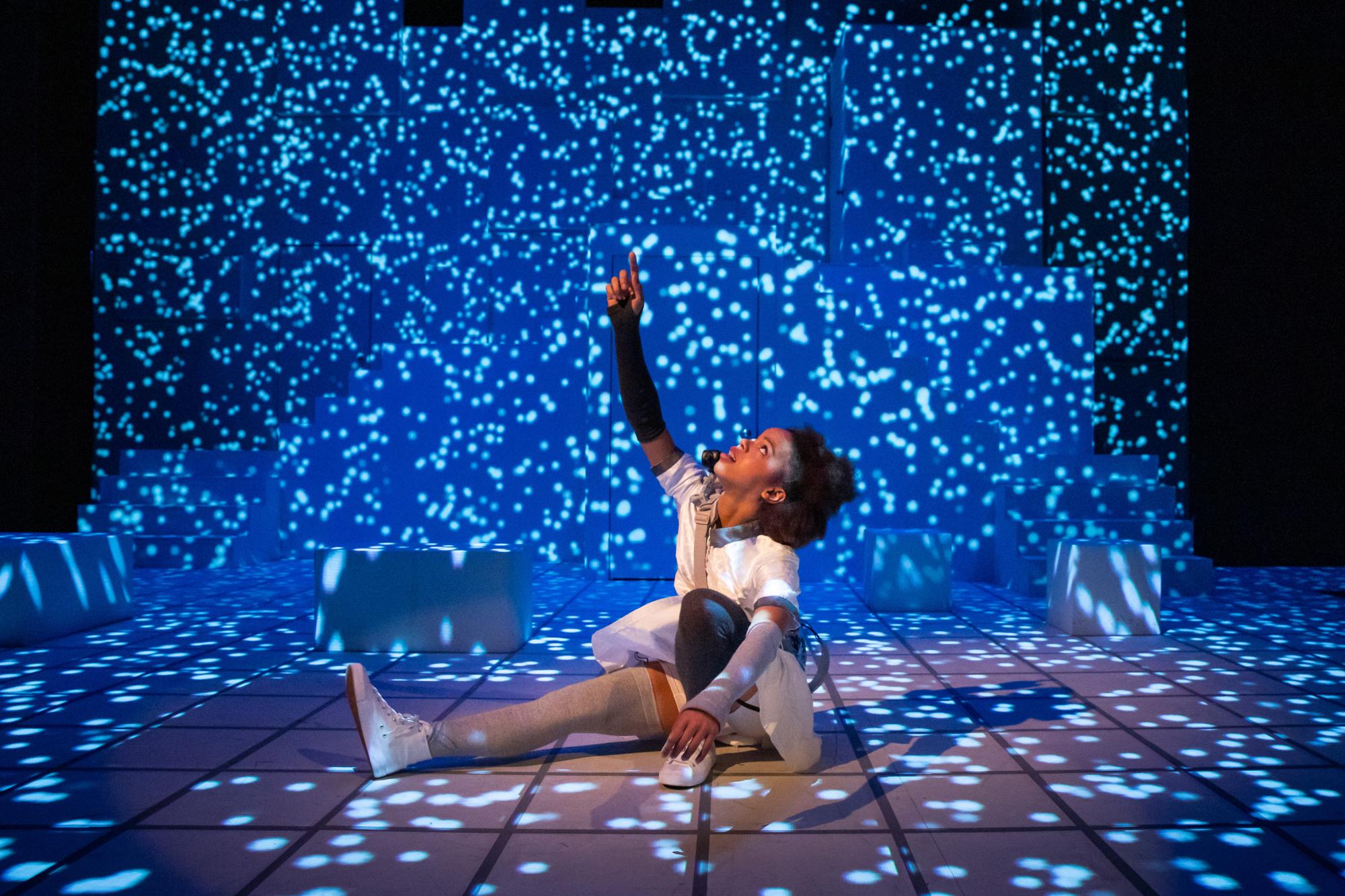actress sits on a stage with hundreds of stars projected on it, lifting her hand and looking up