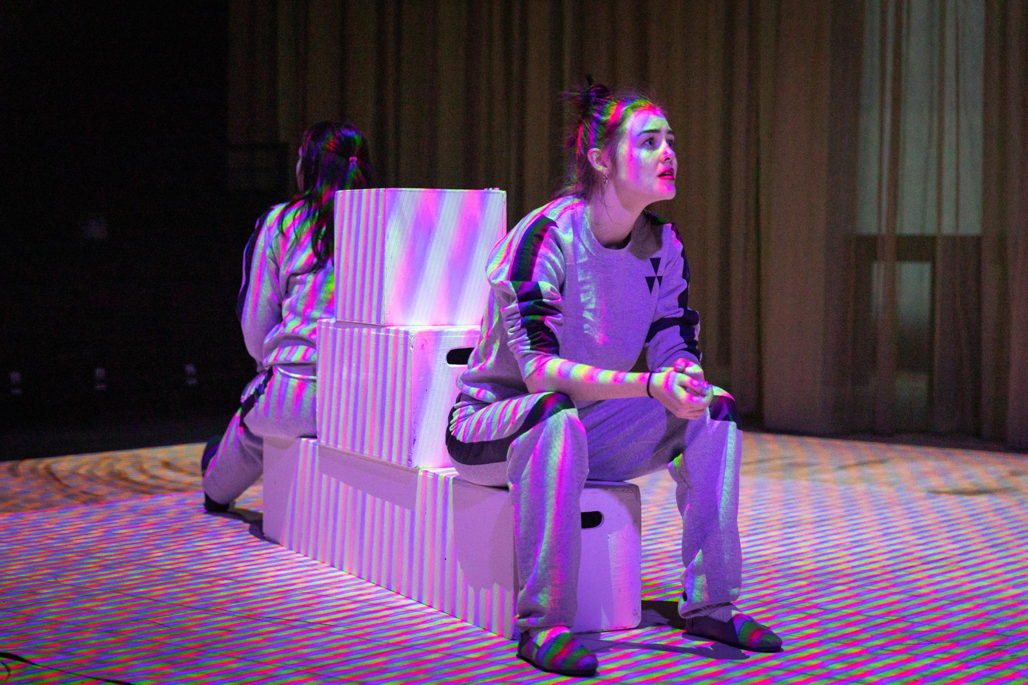 two actors sit on blocks facing opposite directions, checkered with colorful projections