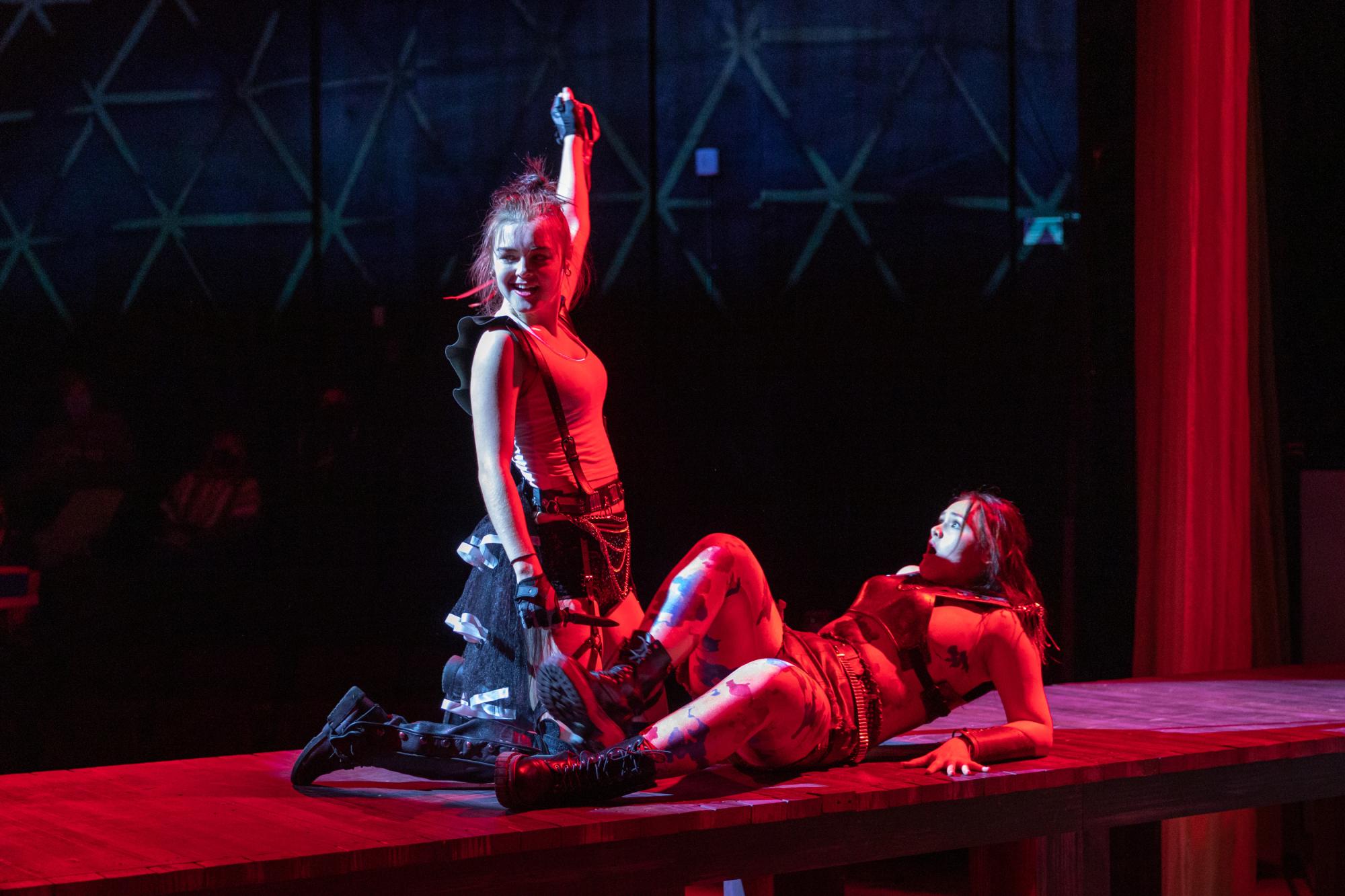 an actor holds up their hand triumphantly, while the actor below them looks horrified, with red stage lighting