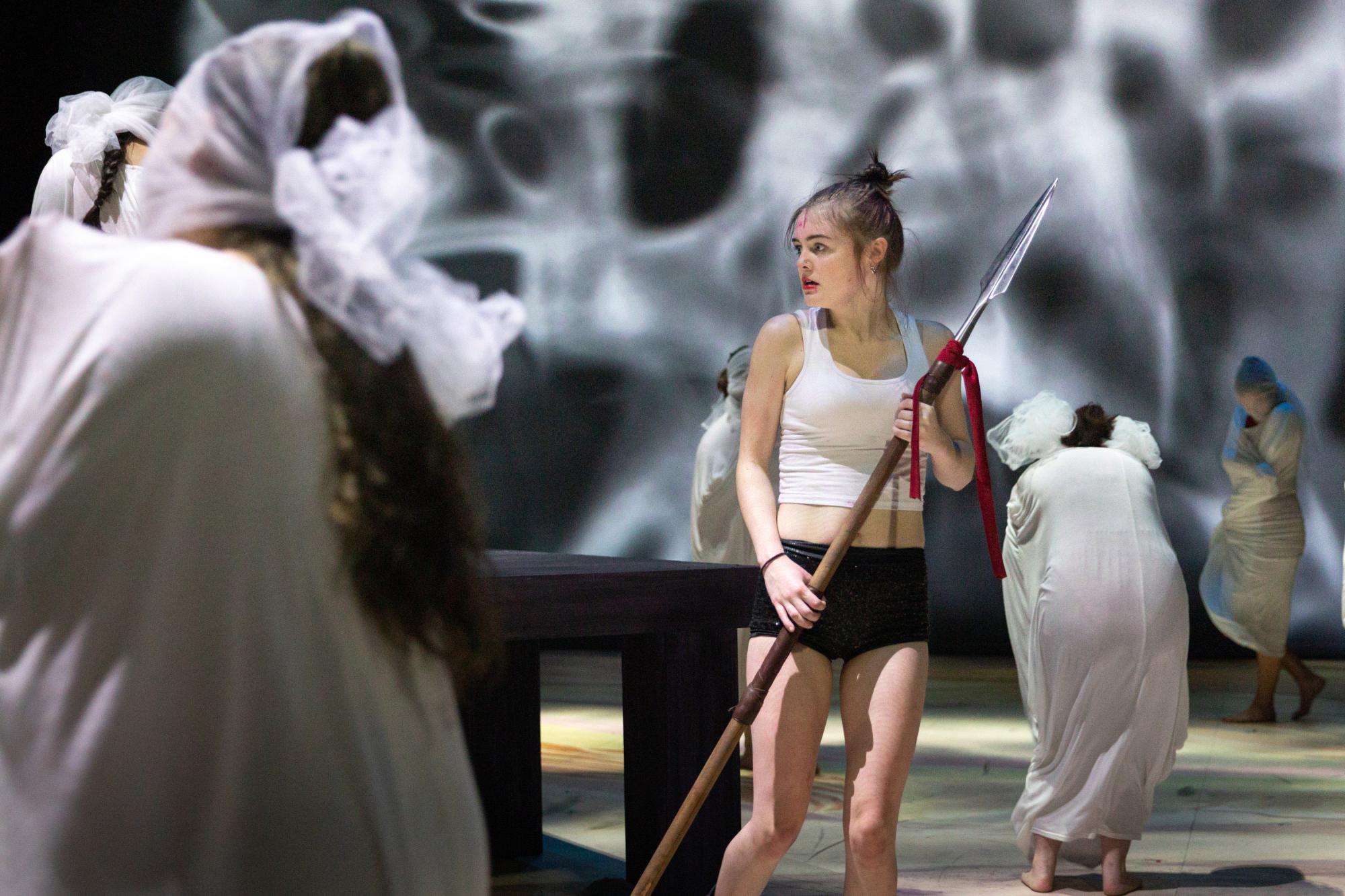 actor holding a spear looks at an ensemble of women cloaked in white