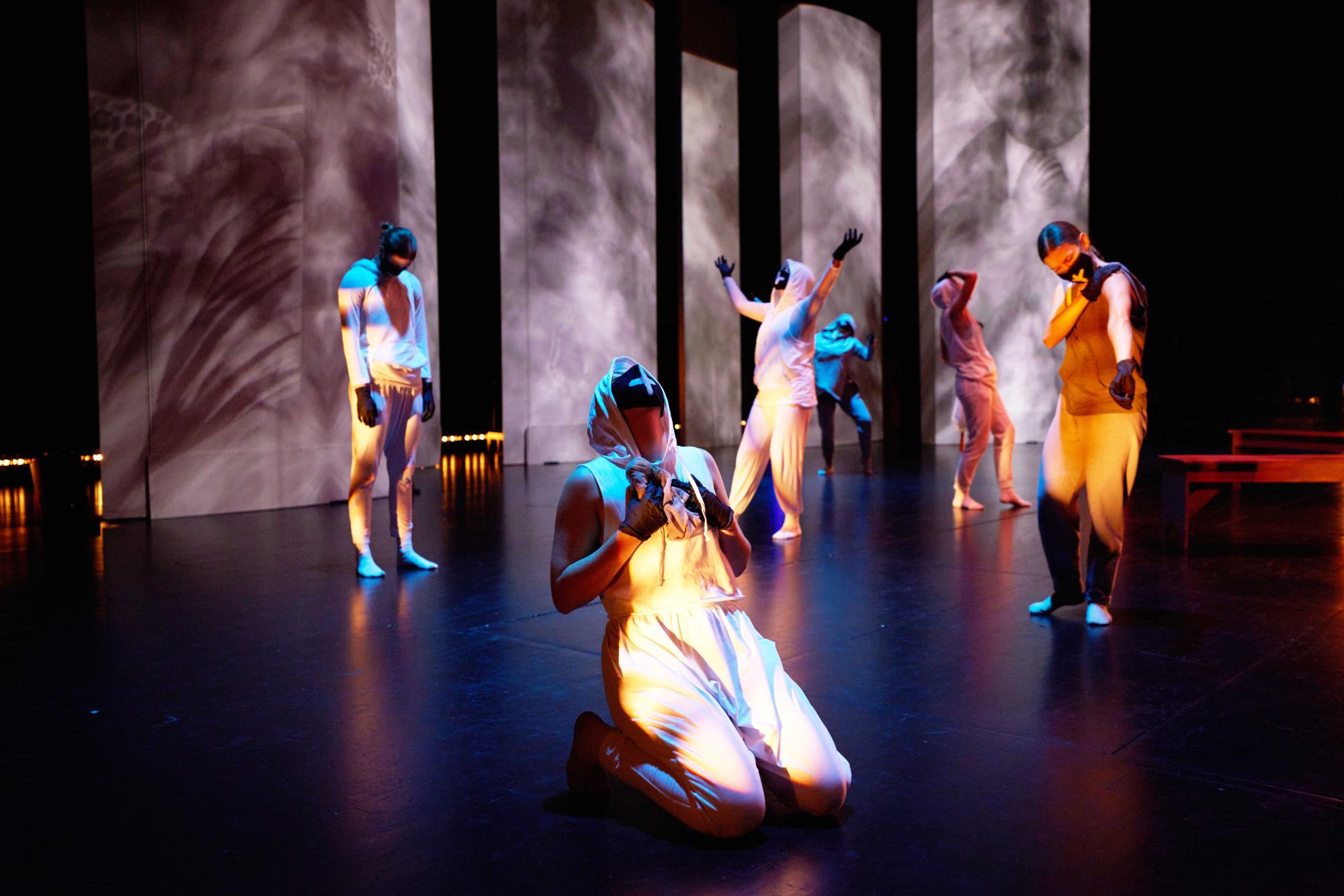 dancer in all white faces upwards, sitting on their knees with five other dancers moving behind them