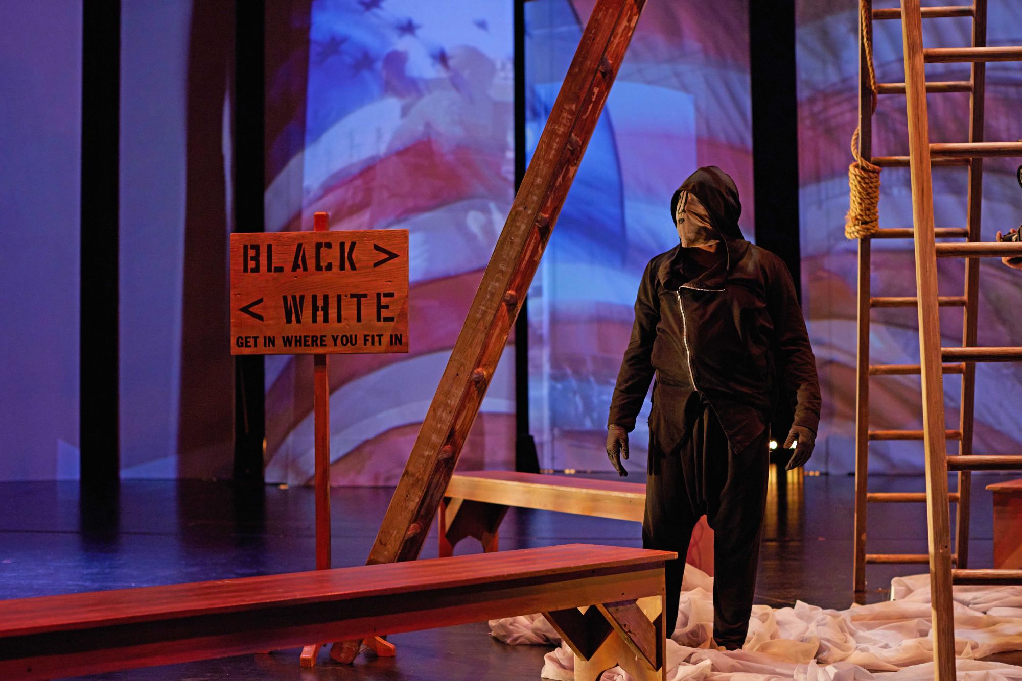 dancer in all black, wearing a full face mask and a hoodie stands beneath a structure of four ladders
