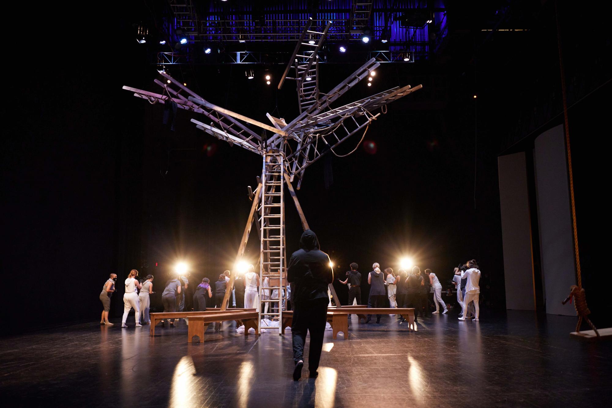 dancer in all black with his hood up walks towards an ensemble of dancers and a tree-like structure made of ladders