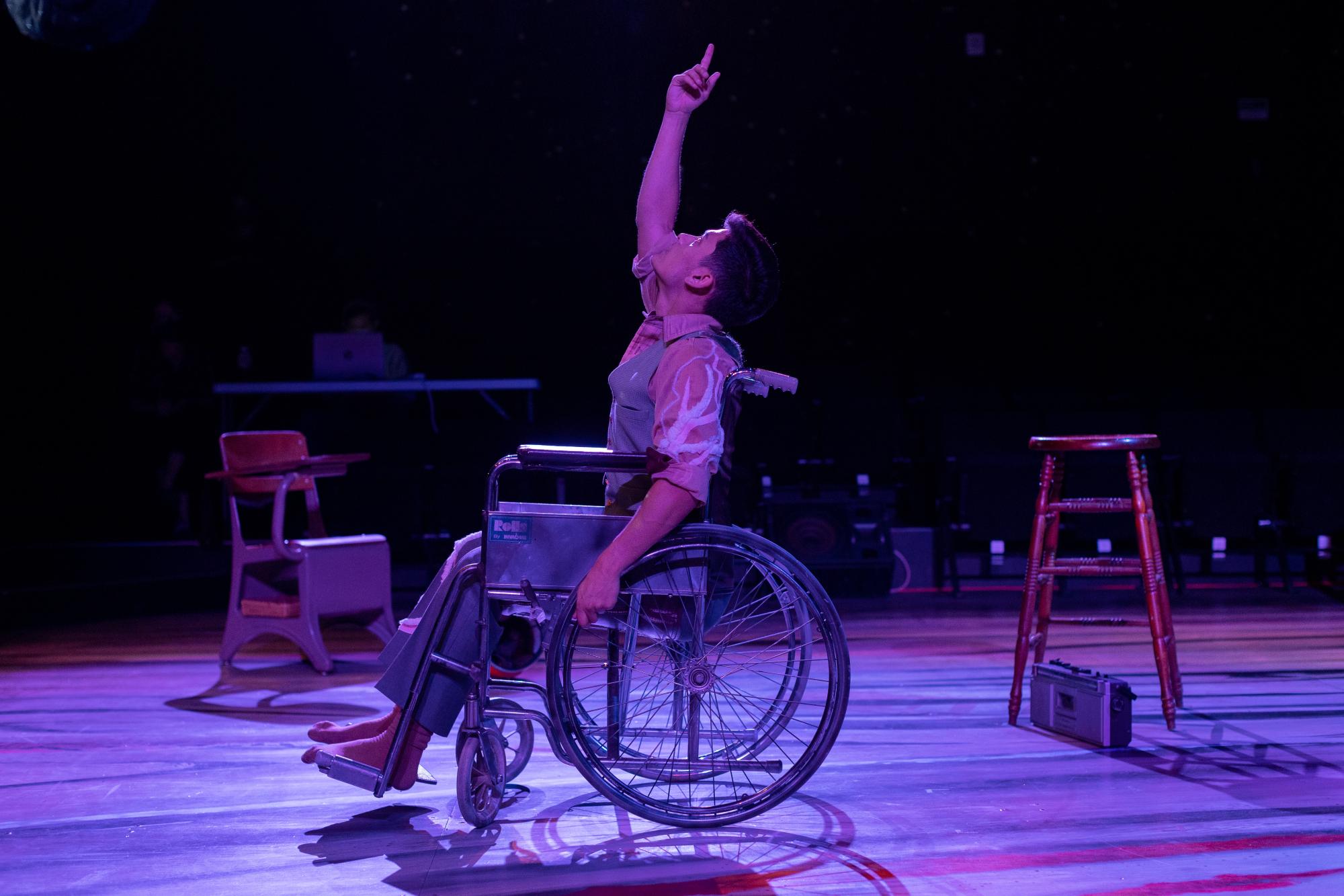 man in a wheelchair points upward, washed in purple stage lighting