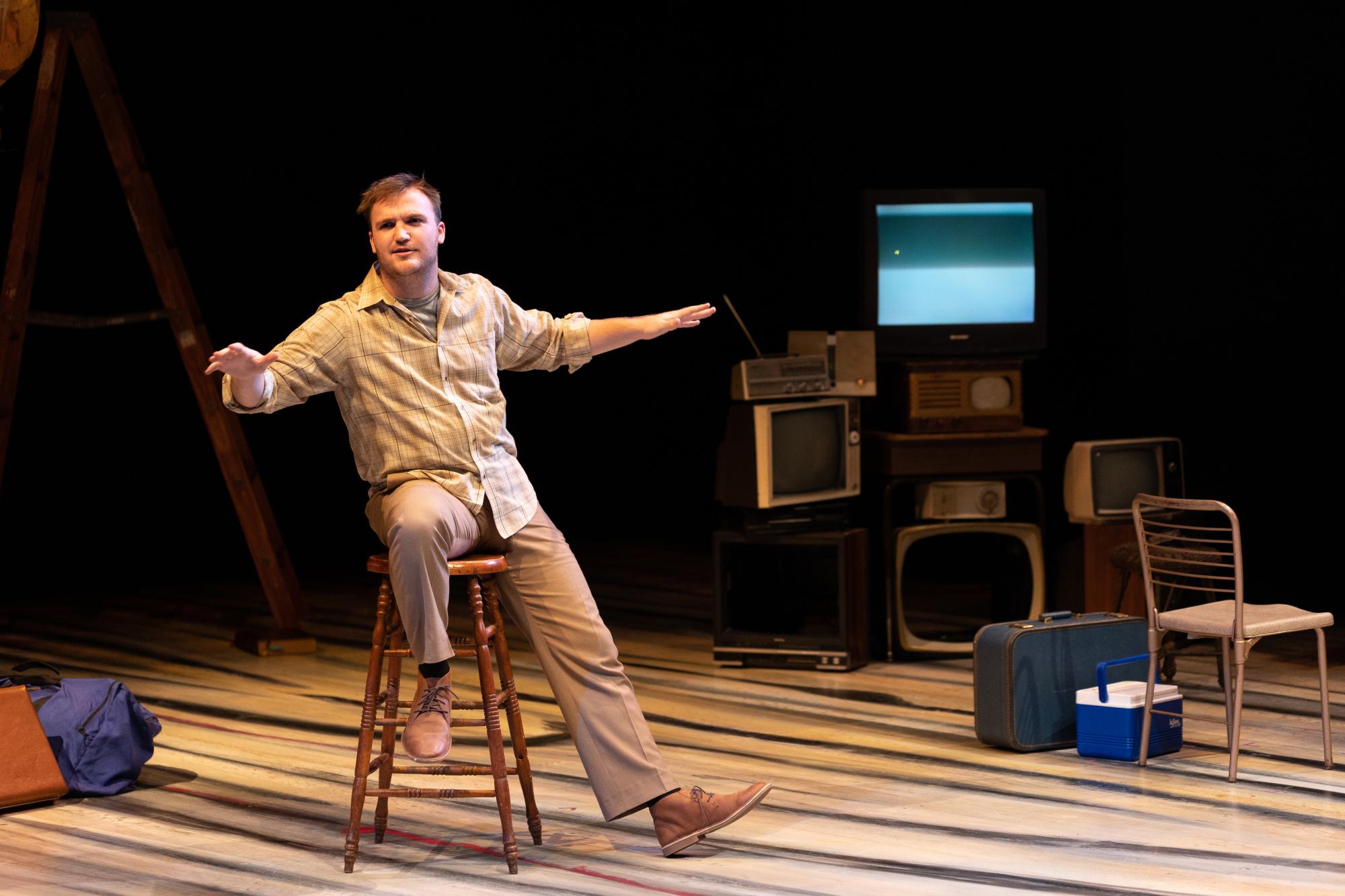 man sits on a stool with his hands outstretched, speaking to an audience of people