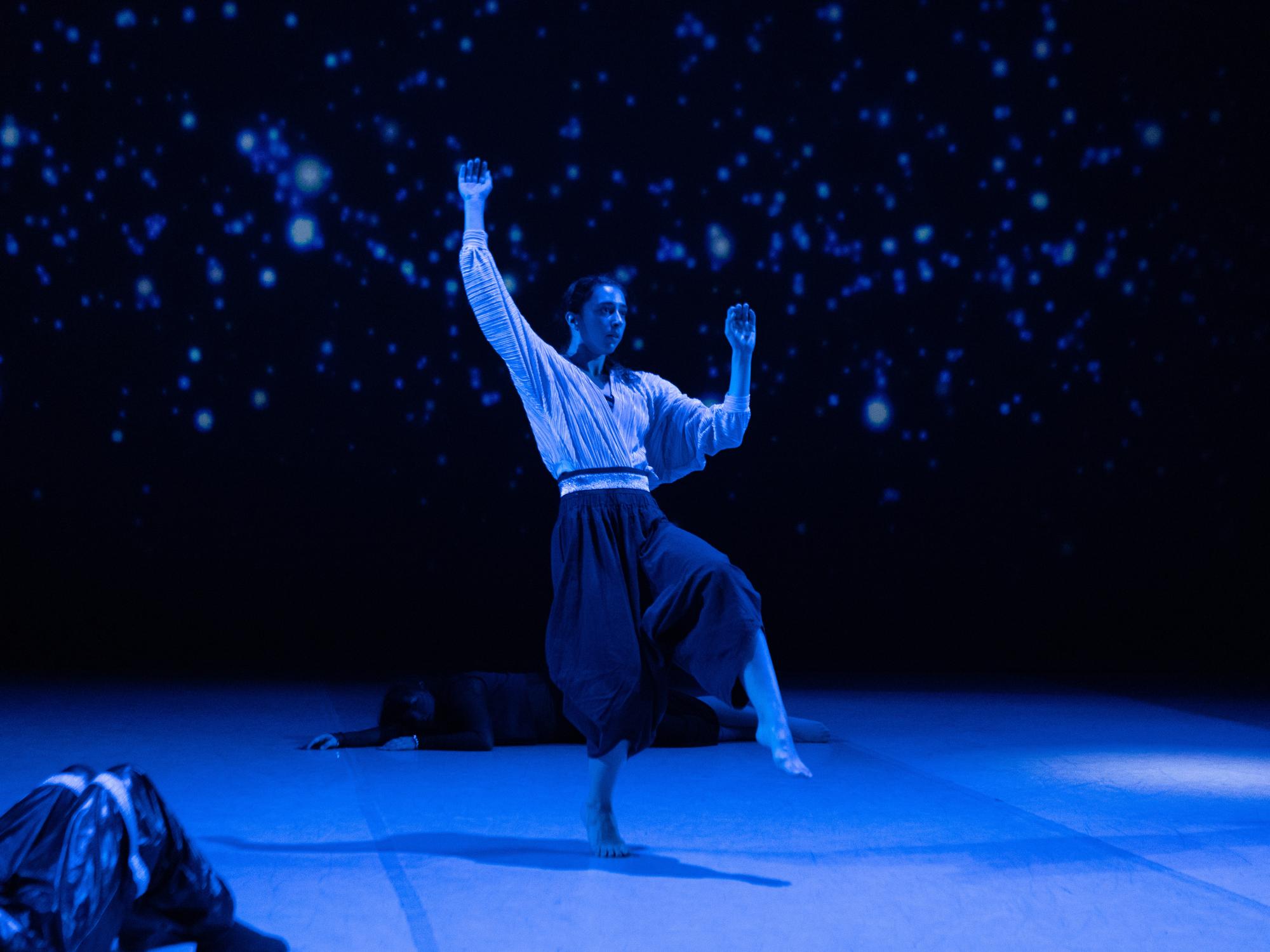 dancer reaches her right arm up and her left leg up slightly, washed in blue light